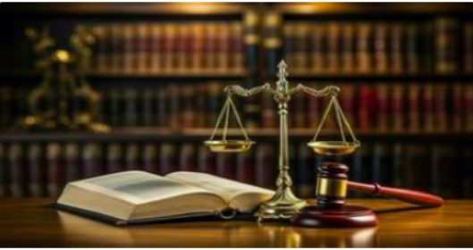 Photo of the scales of justice, a gavel and a book open on a table. A bookshelf with law books is in the background.